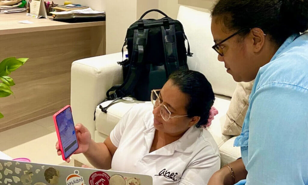 Duas mulheres olham para a tela de um celular ao lado de um laptop aberto.