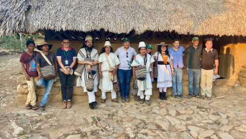 Twelve people standing in front of a thatched roof building. Four of the people are wearing traditional indigenous Colombian clothing.