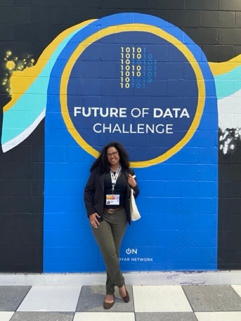 Amaya standing in front of a wall mural that says "Future of Data Challenge"